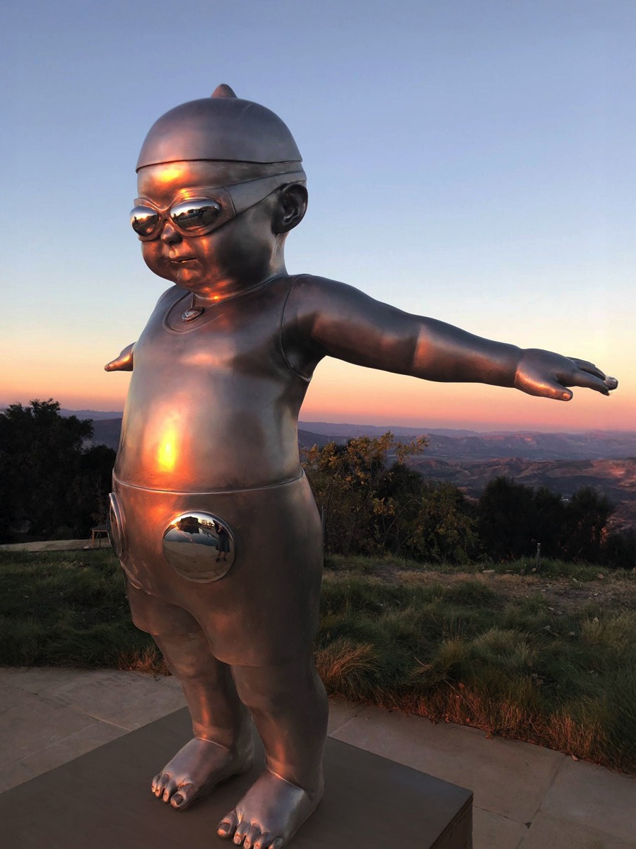 3-4 view of King Tot (Flying) narrative stainless steel figurative sculpture by Richard Becker installed outdoors at sunset