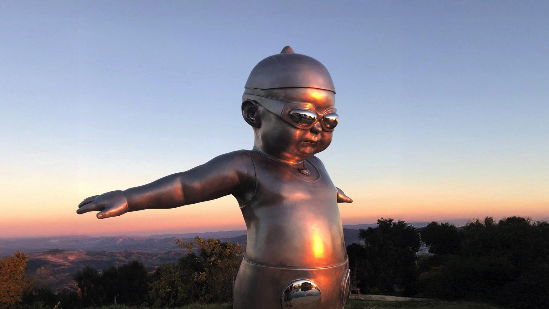 King Tot (Flying) narrative stainless steel figurative sculpture by Richard Becker installed outdoors at sunset